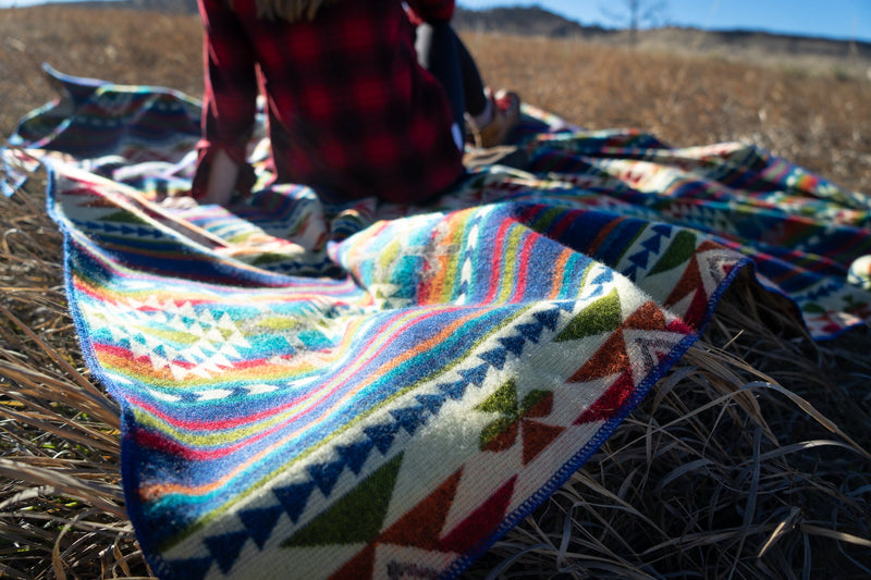 Galapagos Alpaca Blanket spread out loosely on the dry grass, showcasing the vibrancy of the colors against the neutral field.