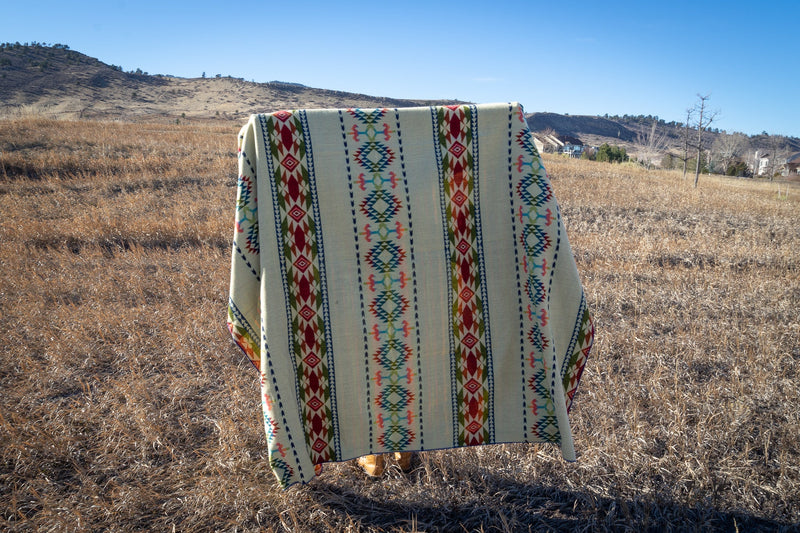 Galapagos Alpaca Blanket held vertically, showcasing the full repeating geometric stripe pattern against a hillside backdrop.