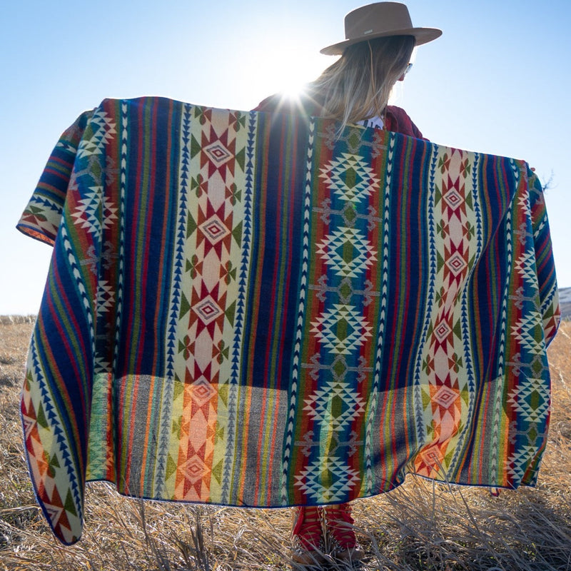 Woman in a hat and red boots holding the Galapagos Alpaca Blanket spread wide, with sun flare highlighting the vibrant geometric stripes in a grassy field.