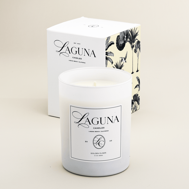 Studio shot of the Laguna Candles Golden Dunes 13oz candle in a white frosted glass vessel next to its white box with black tropical palm illustration.