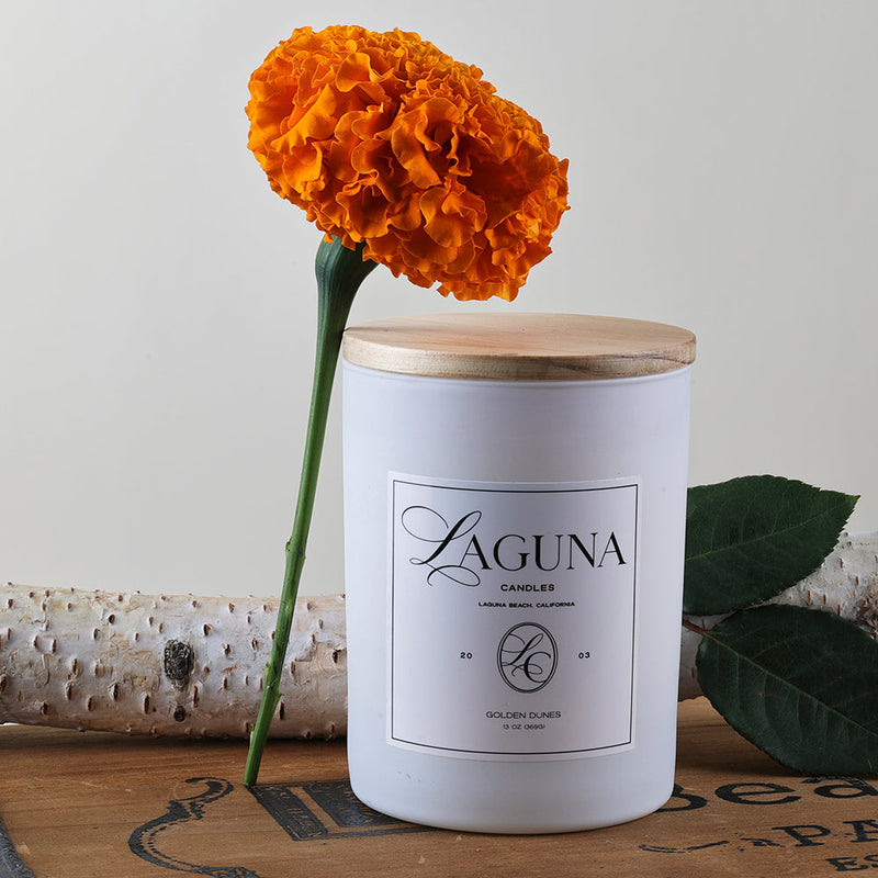 Styled shot of the Golden Dunes 13oz candle with a natural wood lid, featuring an orange marigold flower and birch log accents, suggesting a warm, elegant seasonal ambiance.