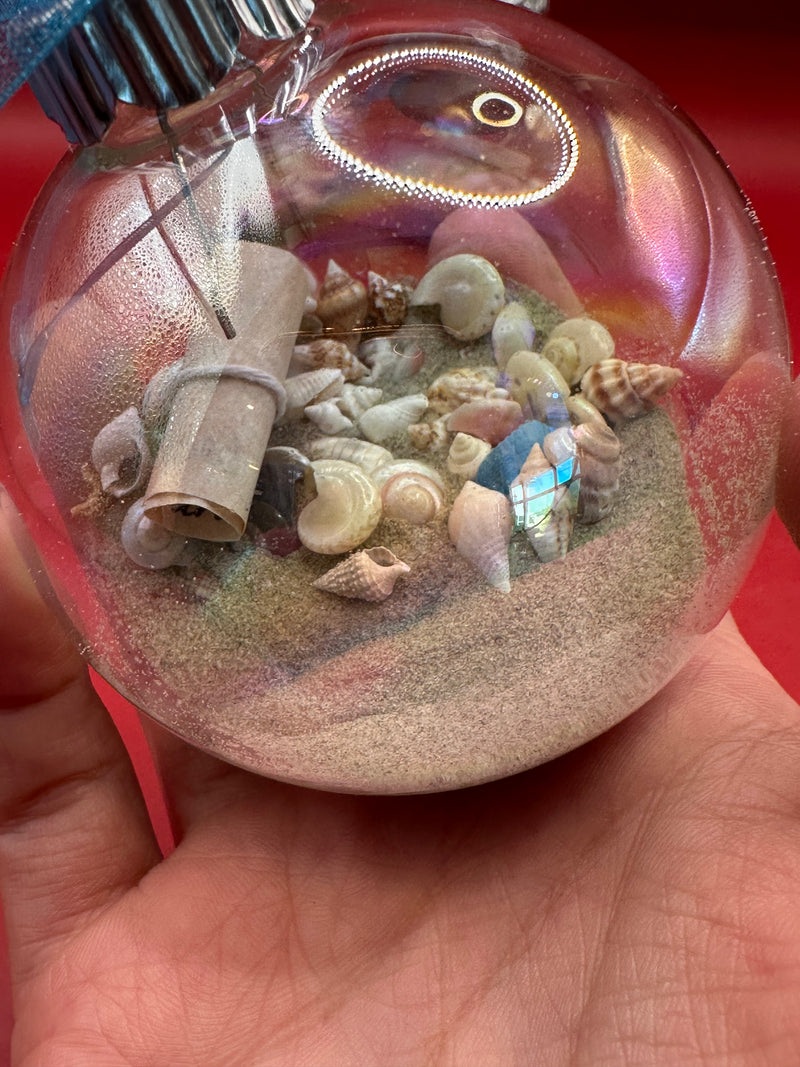 Hand holding coastal glass ornament with sand, assorted mini seashells, and scroll inside for beach Christmas décor