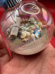 Hand holding coastal glass ornament with sand, assorted mini seashells, and scroll inside for beach Christmas décor