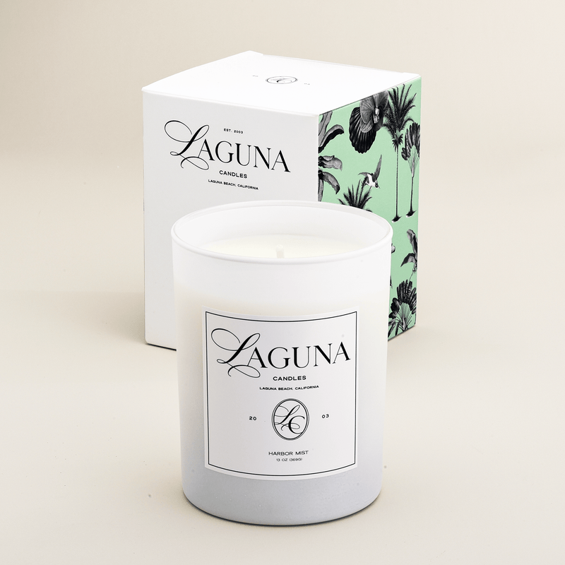Studio shot of the Laguna Candles Harbor Mist 13oz candle in a white frosted vessel next to its white box with mint green and white tropical palm illustration.