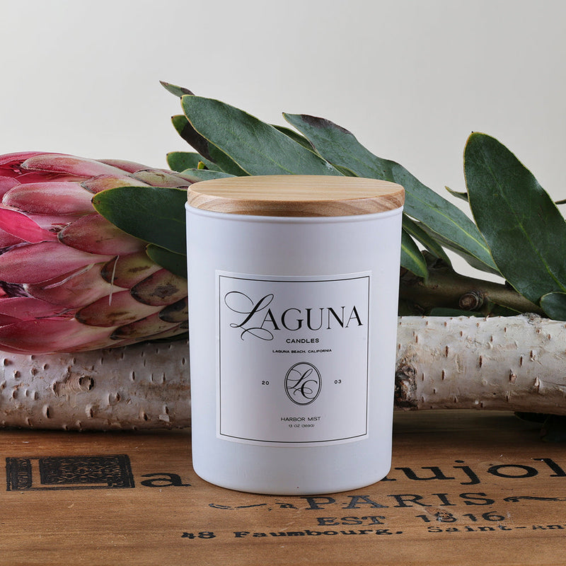 Styled shot of the Harbor Mist 13oz candle with a natural wood lid, accented by a striking pink Protea flower and botanical leaves, emphasizing the sophisticated, complex scent profile.