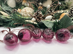 Close-up of five mulberry-colored crackle baubles with ornate caps, placed in front of a festive greenery display, including pinecones and seashells.