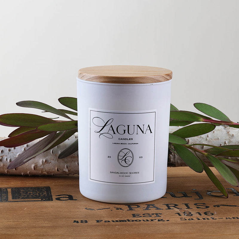 Styled shot of the Laguna 13oz white frosted candle with a natural wood lid, resting on a wooden surface with white linen and green botanical accents.