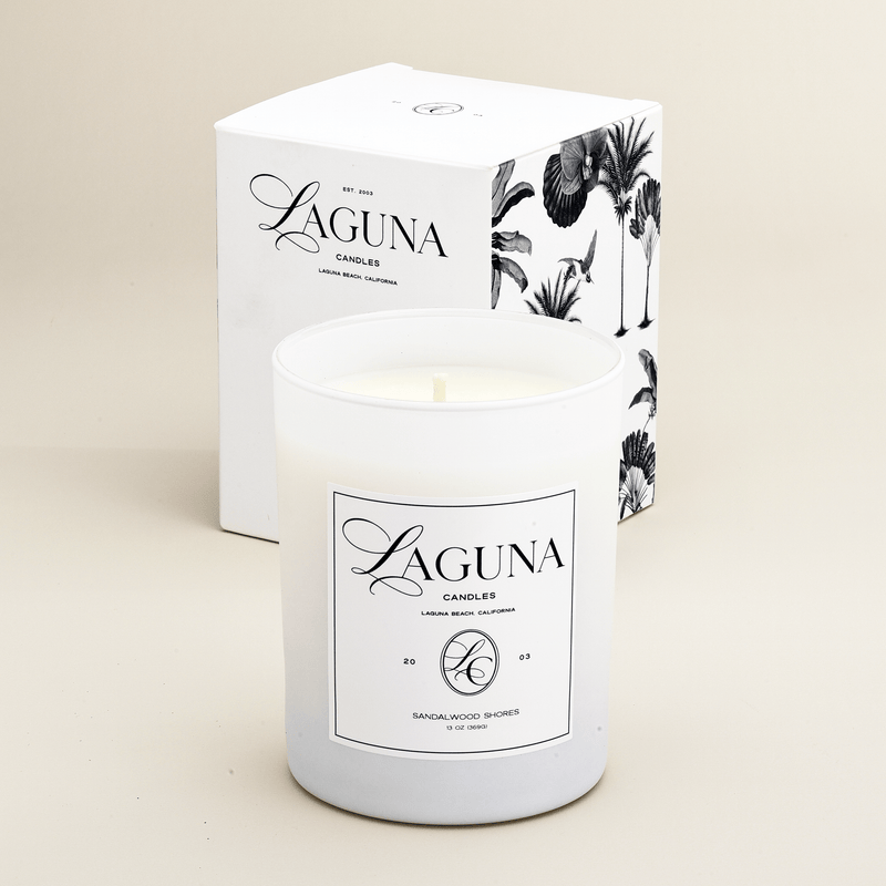 Studio shot of the Laguna 13oz luxury candle in a white frosted glass vessel next to its elegant white gift box with black tropical palm illustrations.