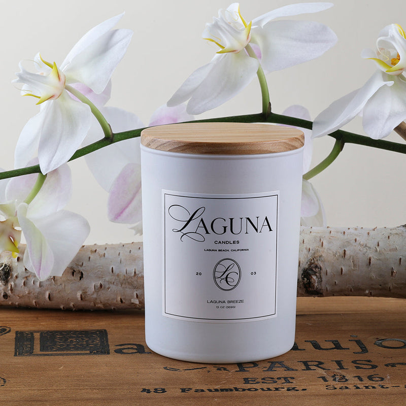 Close-up of a Laguna Candles Laguna Breeze 13oz candle in a white frosted glass vessel with a wooden lid, styled with delicate white orchid flowers in the background, suggesting a serene home ambiance.