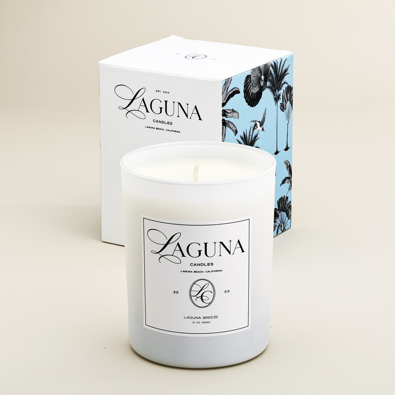 Laguna Candles Laguna Breeze 13oz luxury candle in a frosted white glass vessel, positioned in front of its elegant white gift box adorned with blue tropical palm leaf illustrations.