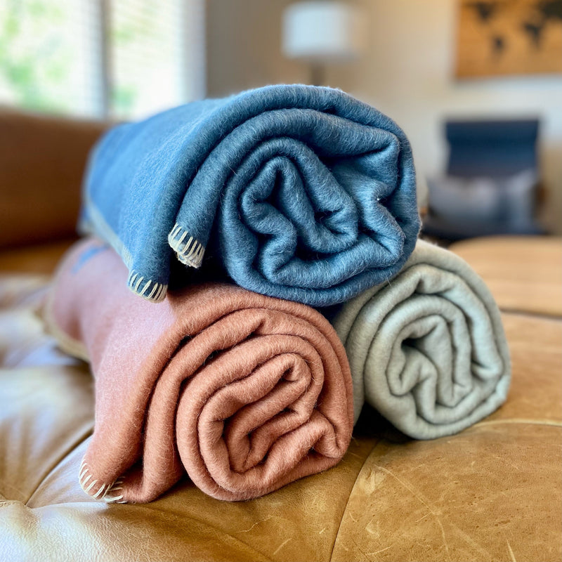 Three tightly rolled luxury wool or alpaca throw blankets stacked on a brown leather surface, showing blue, terracotta/rose, and light gray colors with decorative stitch trim