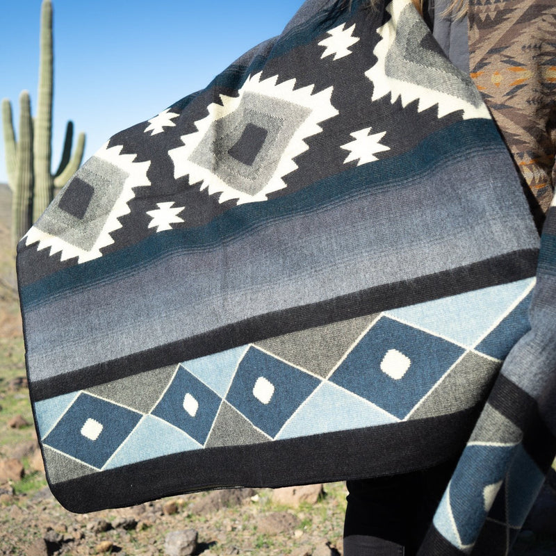 Close-up of the Midnight Blue & Grey Alpaca Blanket draped, showing the dark geometric diamonds, slate stripes, and saguaro cacti in the background.