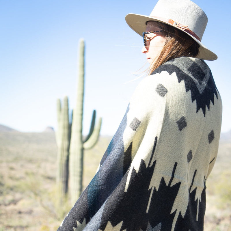 Woman seen in profile, wearing a hat and draped in the Midnight Blue & Grey Blanket, standing in the bright desert sunlight.