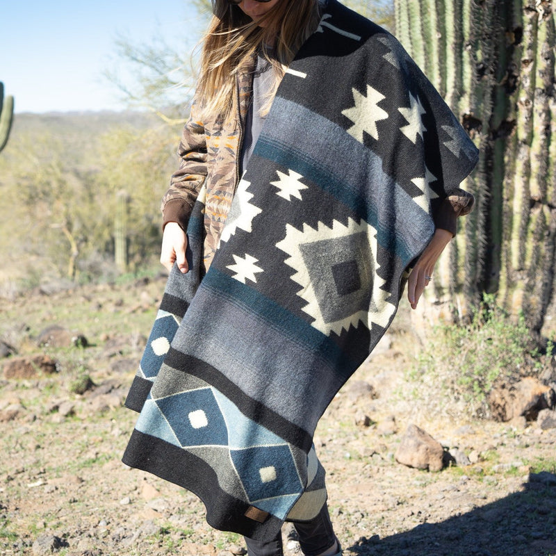 Woman wrapped fully in the Midnight Blue & Grey Blanket, standing outdoors in a desert landscape with saguaro cacti.
