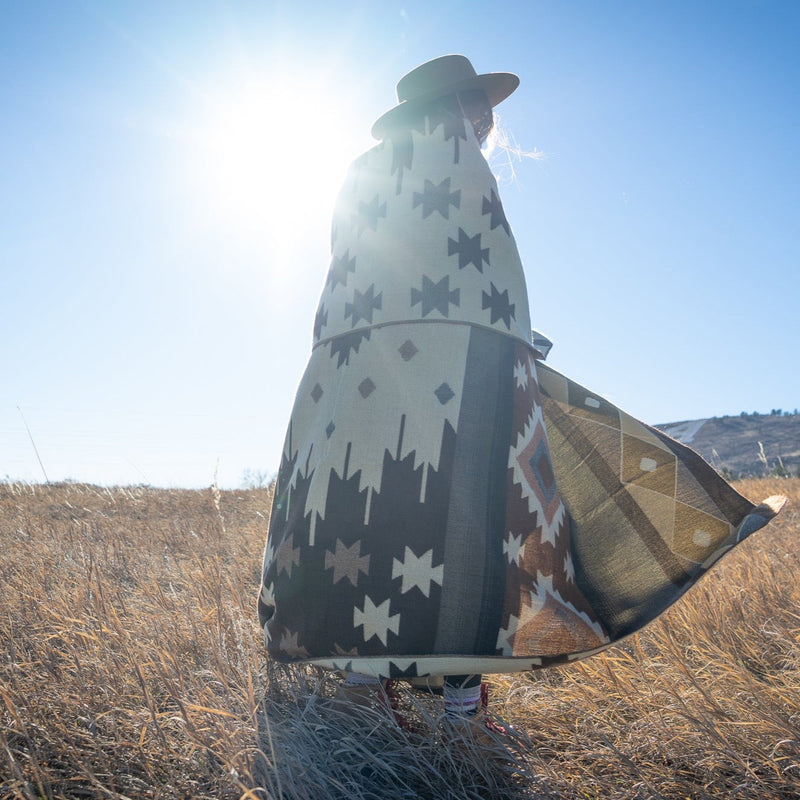 Woman wrapped in the Mojave alpaca blanket, with bright sun flare behind her, emphasizing the warm tones.