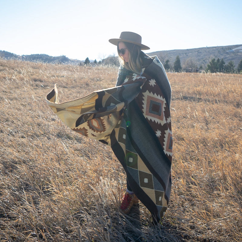 Woman holding the Mojave alpaca blanket draped, standing in a wide, golden grassy field.