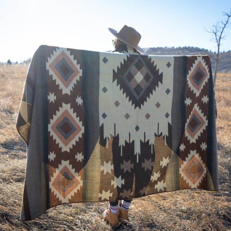 Woman in a hat holding up the Mojave Queen Alpaca Blanket, showcasing the large diamond and stripe geometric pattern in brown and cream earth tones in a grassy field.