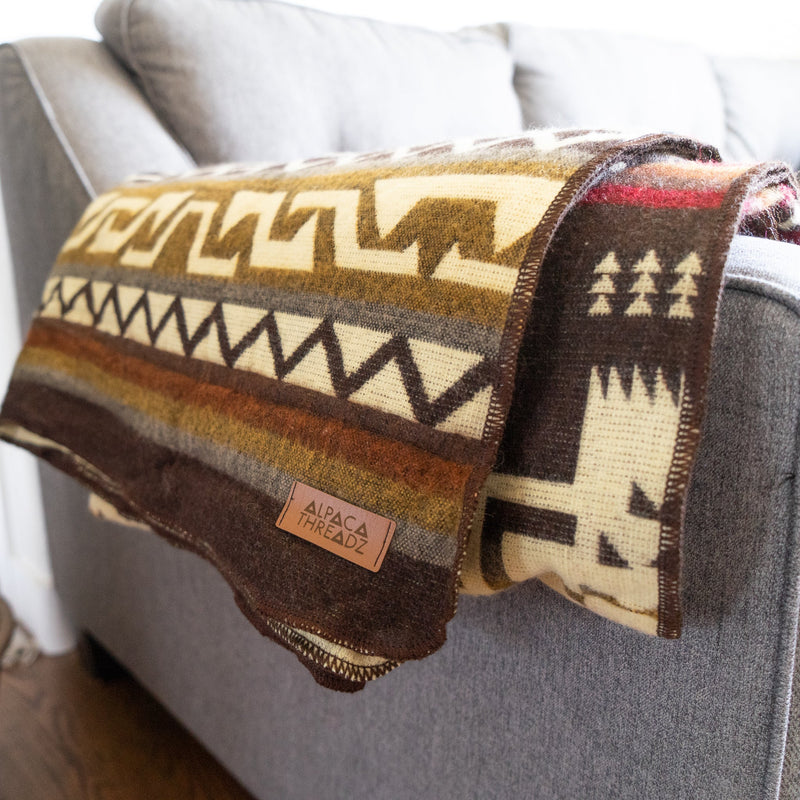 Tightly folded Mountain Alpaca Blanket draped over the corner of a gray sofa, showcasing the layered geometric pattern and label.