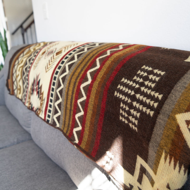 The reversible Mountain Alpaca Blanket draped on a gray sofa, showing the darker, rich brown/burgundy side of the tribal pattern.