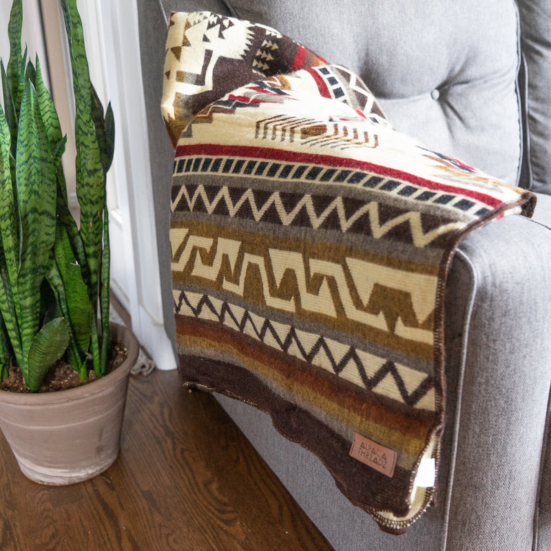 The Mountain Alpaca Blanket draped over a light gray sofa next to a plant, showcasing the cream/light side of the pattern clearly.