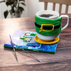 White mug with green and yellow crochet cozy on a book, with a plant in the background