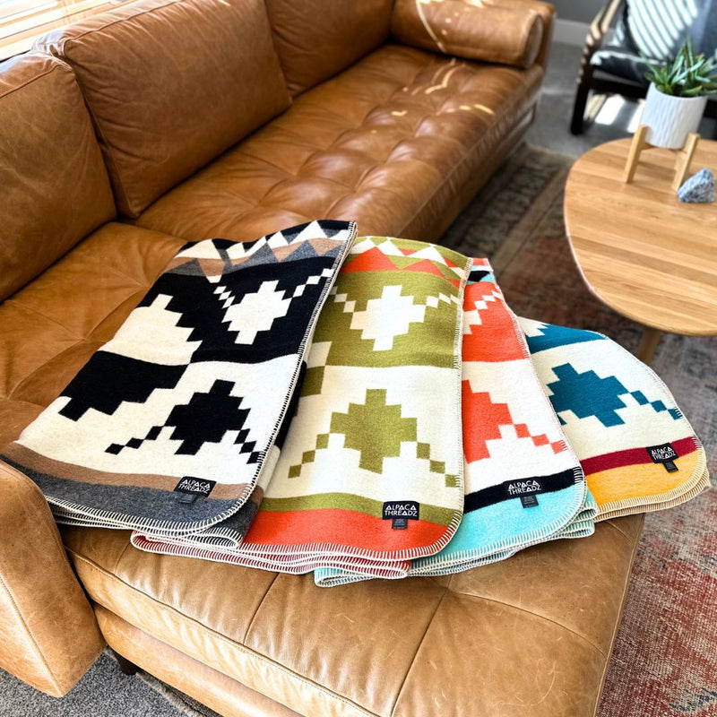 Two multicolor geometric alpaca throws stacked and folded on a brown leather sofa, showcasing the vibrant variants.