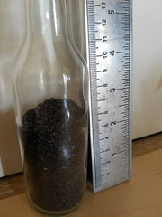 Clear glass bottle filled with dark black sand next to a ruler on a wooden surface
