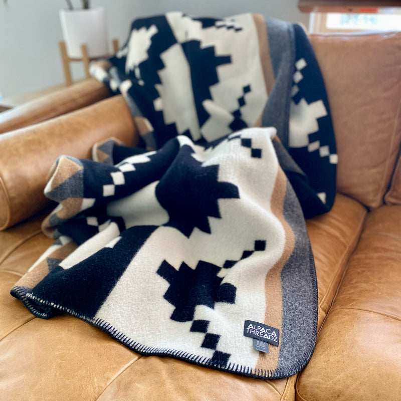 Navy and white geometric alpaca throw blanket folded and draped over the corner of a soft gray or brown sectional sofa.