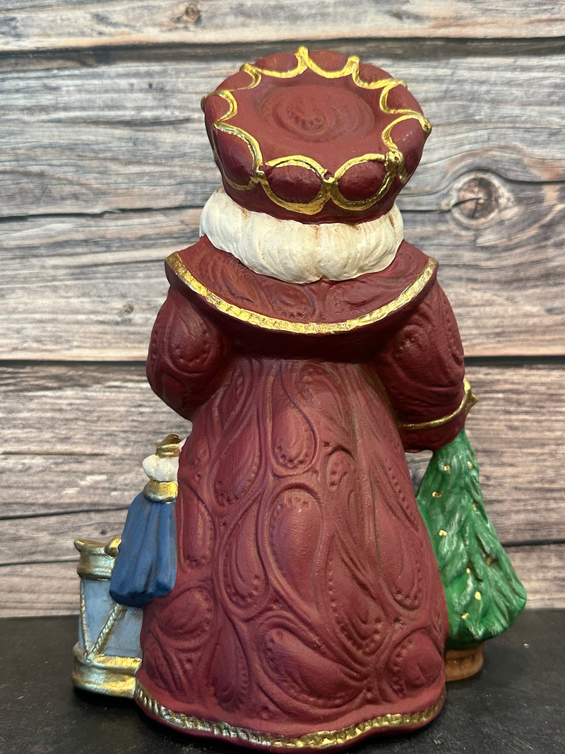 Ceramic Santa figurine in a burgundy robe, holding a nutcracker and ballerina, surrounded by ballet slippers, toy soldiers, and a Christmas tree, with intricate gold accents.