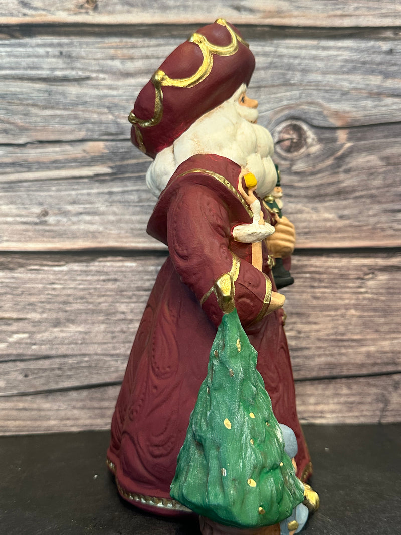 Ceramic Santa figurine in a burgundy robe, holding a nutcracker and ballerina, surrounded by ballet slippers, toy soldiers, and a Christmas tree, with intricate gold accents.