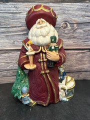 Ceramic Santa figurine in a burgundy robe, holding a nutcracker and ballerina, surrounded by ballet slippers, toy soldiers, and a Christmas tree, with intricate gold accents.