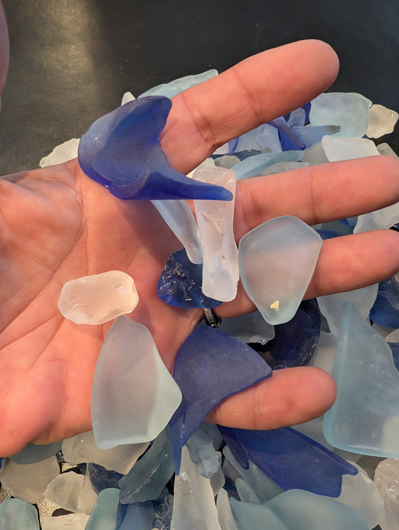 A hand holding assorted pieces of frosted sea glass in blue and clear hues from the Ocean Blue Mix.