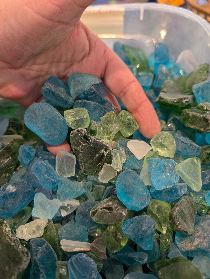 A hand holding pieces of hand-tumbled sea glass in a mix of blue, green, and aqua, showcasing the frosted texture and unique shapes of the glass.