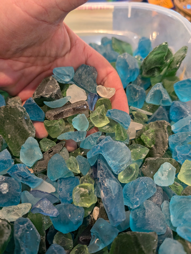 A hand sifting through a collection of hand-tumbled sea glass in blue, green, and aqua, displaying the variation in colors and sizes of the frosted glass pieces.