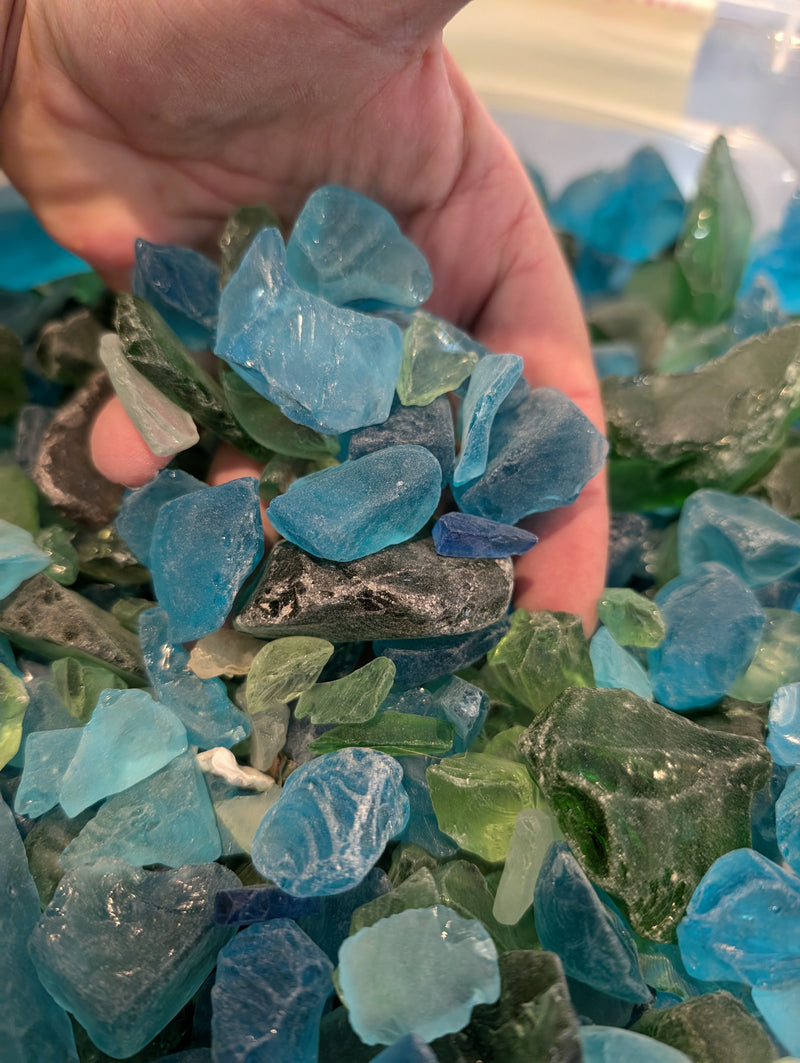 A hand sifting through a collection of hand-tumbled sea glass in blue, green, and aqua, displaying the variation in colors and sizes of the frosted glass pieces.