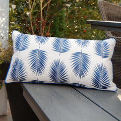 Palm Pattern Indoor/Outdoor Pillow featuring embroidered blue palm leaves on a white background with blue piping, displayed on a bench.