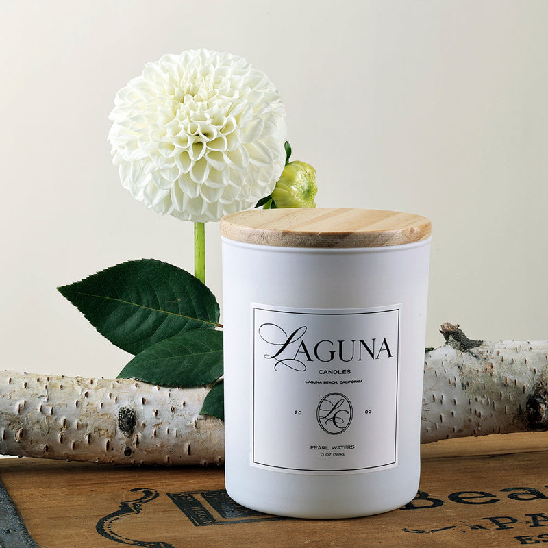 Close-up of the Pearl Waters 13oz candle with a wooden lid, styled against birch logs and a white dahlia flower, highlighting the clean, rustic elegance.