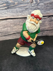 Santa figurine playing pickleball, wearing a green shirt, red shorts, and a red headband, holding a paddle with a yellow pickleball, standing on a white base with a wooden background.