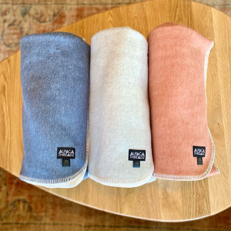 Three rolled Alpaca Throws in blue, white/natural, and rose/pink, with blanket stitch edging and 'Alpaca Threadz' woven labels, displayed on a light wooden table