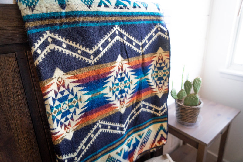 Rio Alpaca Blanket draped on a bed with a wooden side table visible, highlighting the blue and cream geometric pattern.