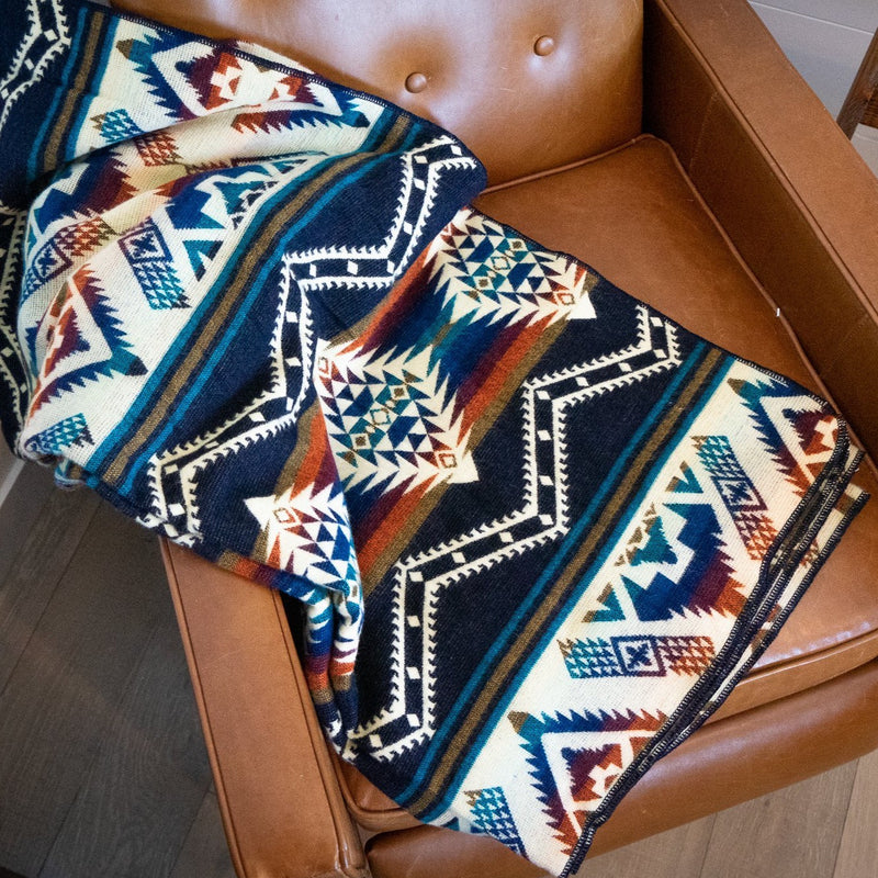 Rio Alpaca Blanket draped over a brown leather armchair, showcasing the vibrant geometric pattern in navy, white, terracotta, and blue.