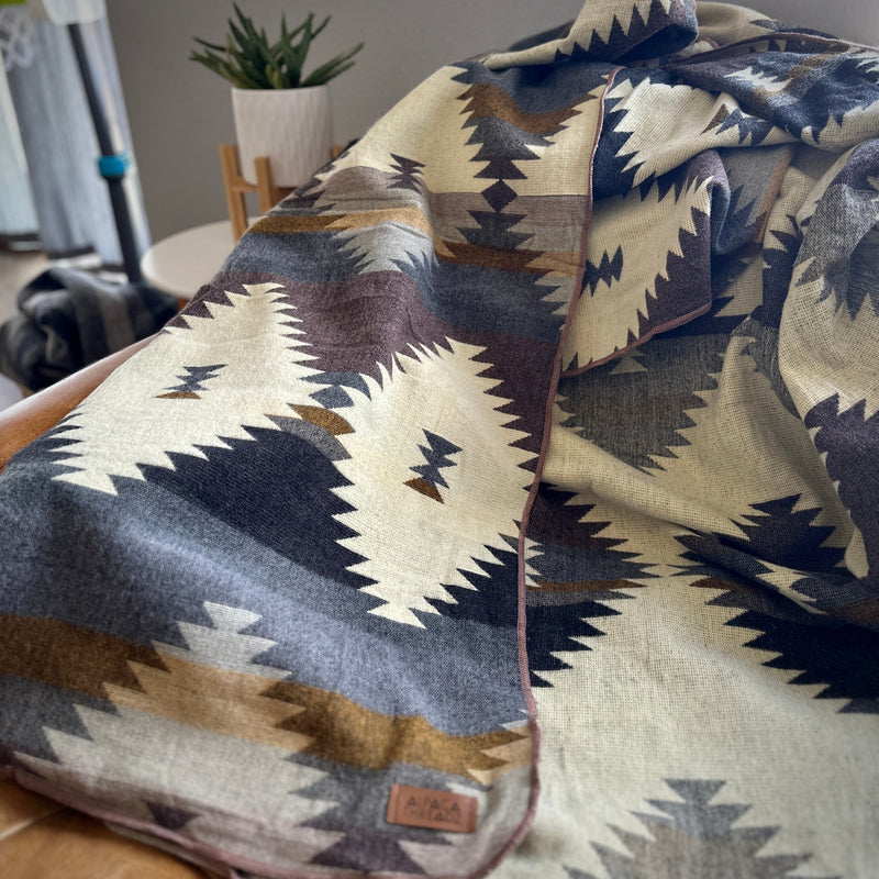 Rocky Alpaca Blanket draped at an angle over a dark armchair, highlighting the contrast between the cream, slate, and dark brown motifs.