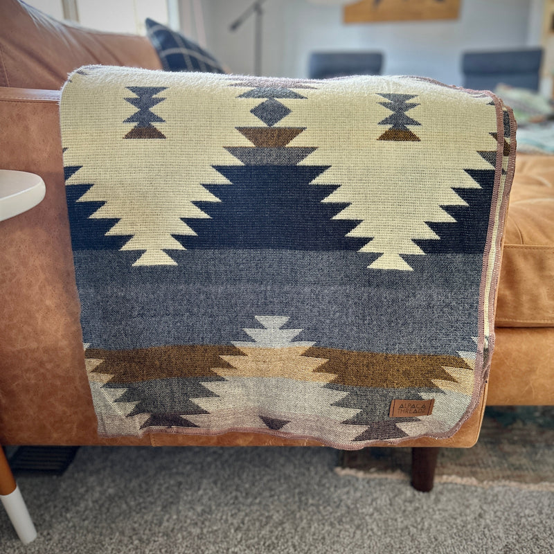 Rocky Alpaca Blanket folded into a square, draped over the back of a light gray or tan sofa, emphasizing the blue and gray geometric pattern.