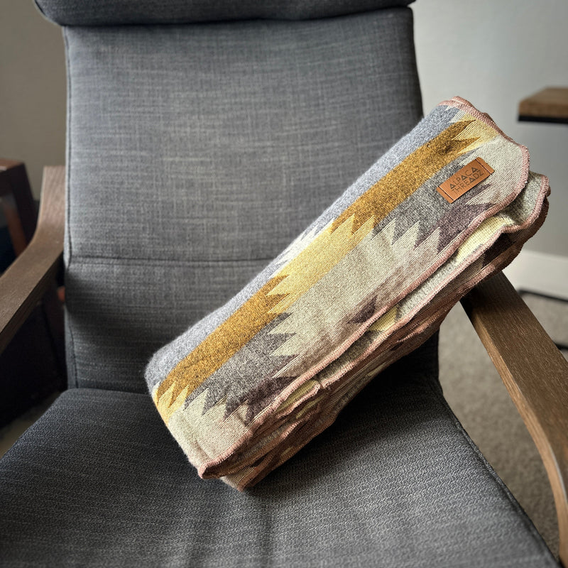 Rocky Alpaca Blanket neatly folded into a stack on a wooden side table next to a gray chair, showcasing the side profile and folded texture.