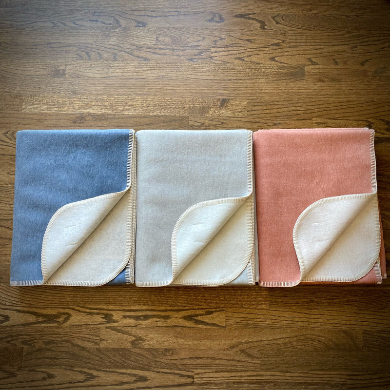 Set of three woven Camp Throw Blankets in salmon pink, sky blue, and light grey colors displayed together on rustic wooden surface, soft textured fabric showing cozy coastal and cabin-inspired style