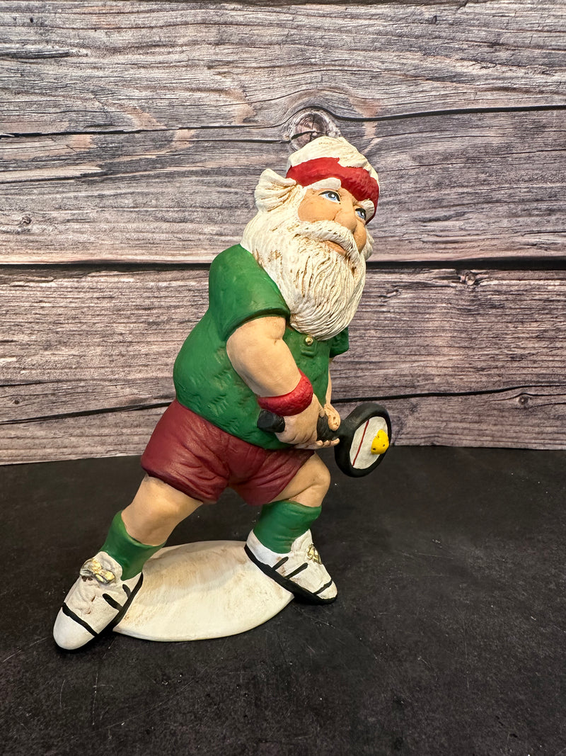 Santa figurine playing pickleball, wearing a green shirt, red shorts, and a red headband, holding a paddle with a yellow pickleball, standing on a white base with a wooden background.Santa figurine playing pickleball, wearing a green shirt, red shorts, and a red headband, holding a paddle with a yellow pickleball, standing on a white base with a wooden background.