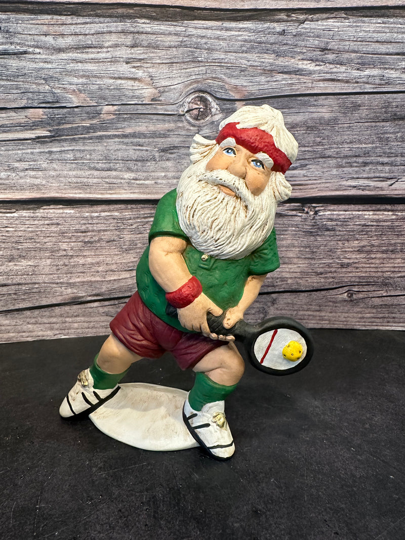 Santa figurine playing pickleball, wearing a green shirt, red shorts, and a red headband, holding a paddle with a yellow pickleball, standing on a white base with a wooden background.