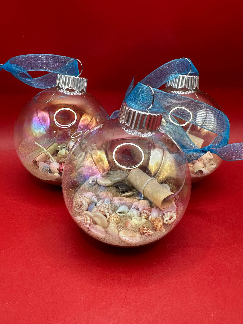Close-up of glass Christmas ornament filled with real sand, tiny seashells, and a rolled message in a bottle tied with turquoise ribbon