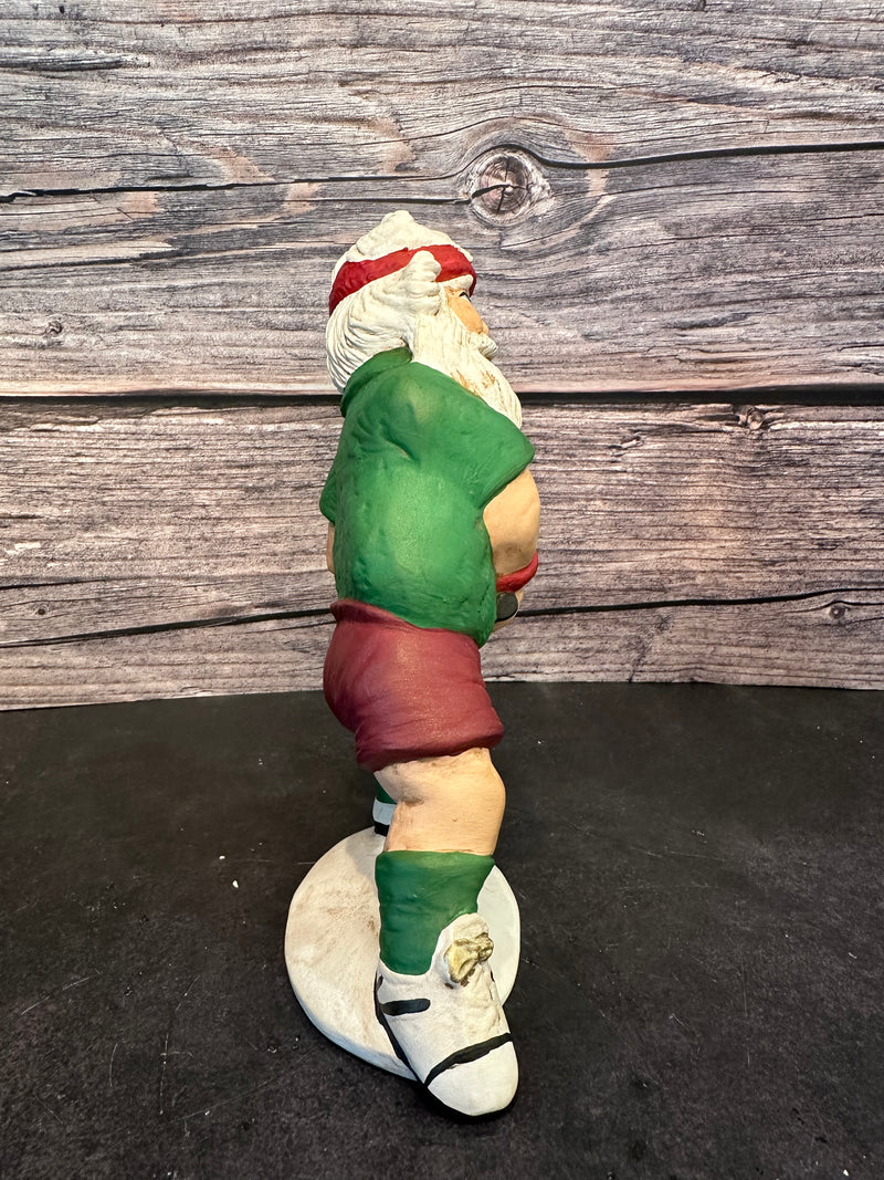 Santa figurine playing pickleball, wearing a green shirt, red shorts, and a red headband, holding a paddle with a yellow pickleball, standing on a white base with a wooden background.