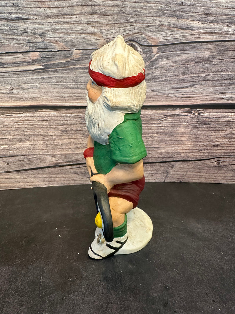 Santa figurine playing pickleball, wearing a green shirt, red shorts, and a red headband, holding a paddle with a yellow pickleball, standing on a white base with a wooden background.